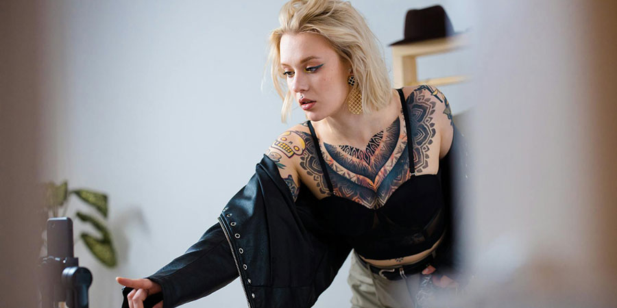 woman with short blonde hair and body tattoo wearing a black jacket in front of a smartphone